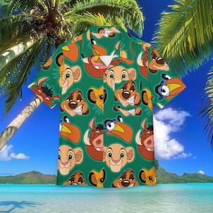 Puede incluir: Camisa verde con botones con un patrón repetido de caras de personajes de dibujos animados en marrón, beige y naranja. La camisa tiene mangas cortas y un cuello clásico. El fondo es una escena tropical con palmeras y cielo azul.