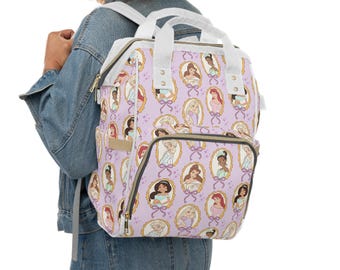 Puede incluir: Mochila de color lila claro con un patrón repetido de retratos de princesas enmarcados. La mochila tiene correas y asas blancas, y múltiples bolsillos. El diseño incluye marcos dorados y lazos morados.