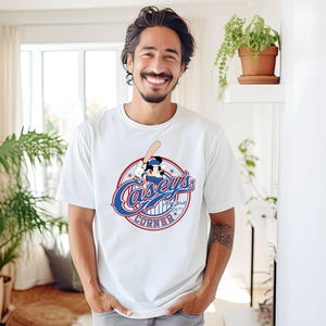 May include: White t-shirt featuring a baseball-themed graphic with the text "Casey's Corner" in a circular design. The design includes a baseball bat and a cartoon character. The shirt is worn by a person.
