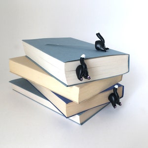 May include: A stack of three books with black cat bookmarks. The bookmarks are shaped like cats with white tails and pink noses. The books are blue, beige, and blue.