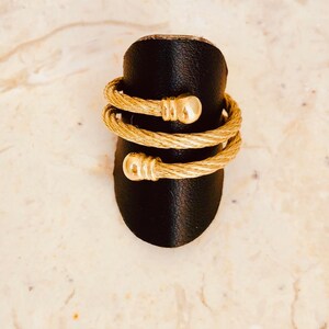 May include: A gold-toned ring with a twisted cable design and rounded ends. The ring is displayed on a black ring holder.