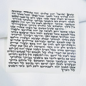 Może przedstawiać: Biały materiał z nadrukowanym czarnym tekstem hebrajskim. Tekst zawiera słowa "Szema Jisrael" i "Adonai Eloheinu Adonai Echad".