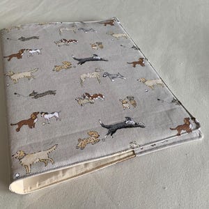 May include: A rectangular fabric book cover in a light grey colour featuring a repeating pattern of various dog breeds in shades of brown, white, black, and tan. The cover has visible stitched edges and a plain cream-coloured interior lining.