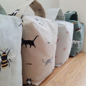 May include: Four fabric doorstops with handles, each with a different pattern. The patterns include a bee, black cats, fish bones, pink hearts, and a Dalmatian dog. The doorstops are in shades of beige, light blue, and green.