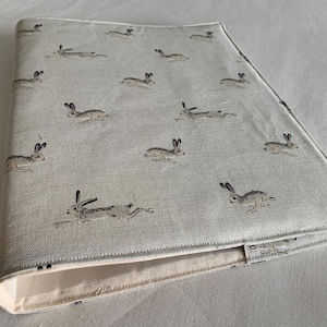 May include: A light beige book cover with a repeating pattern of running hares in grey. The cover has a folded edge and a cream-coloured interior lining. The design is reminiscent of a countryside theme, perfect for nature lovers.