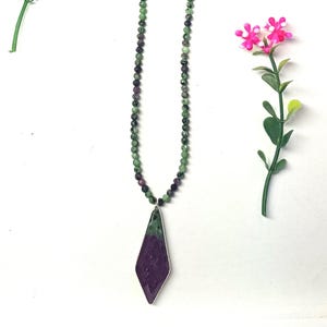 May include: A beaded necklace with a diamond-shaped pendant. The necklace features small, faceted green and purple beads. The pendant is a combination of green and purple stone, set in a silver-colored metal frame. The necklace is displayed on a white surface.