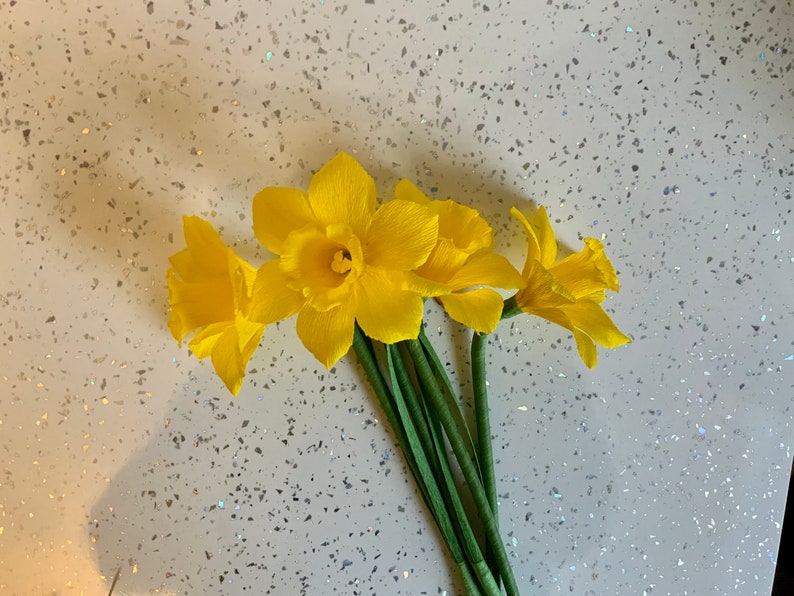 May include: A bouquet of three bright yellow daffodils with green stems. The flowers are in full bloom, with ruffled petals and a prominent central trumpet. The arrangement is set against a speckled white background.