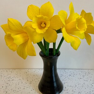 May include: A bouquet of bright yellow paper daffodils in a black vase. The daffodils have ruffled petals and green stems. The vase is a dark, glossy black and sits on a white speckled surface.