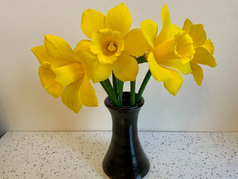 May include: A bouquet of bright yellow paper daffodils in a black vase. The daffodils have ruffled petals and green stems. The vase is a dark, glossy black and sits on a white speckled surface.