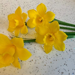 May include: Four bright yellow paper daffodils with green stems are arranged on a speckled white surface. The flowers have ruffled petals and trumpet-shaped centers, showcasing a handmade floral craft.