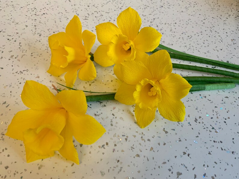 May include: Four bright yellow paper daffodils with green stems are arranged on a speckled white surface. The flowers have ruffled petals and trumpet-shaped centers, showcasing a handmade floral craft.