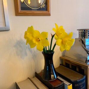 May include: A black ceramic vase holds several bright yellow daffodils. The vase sits atop a stack of books, with a framed photo and a decorative metal lantern in the background. The flowers are in full bloom.