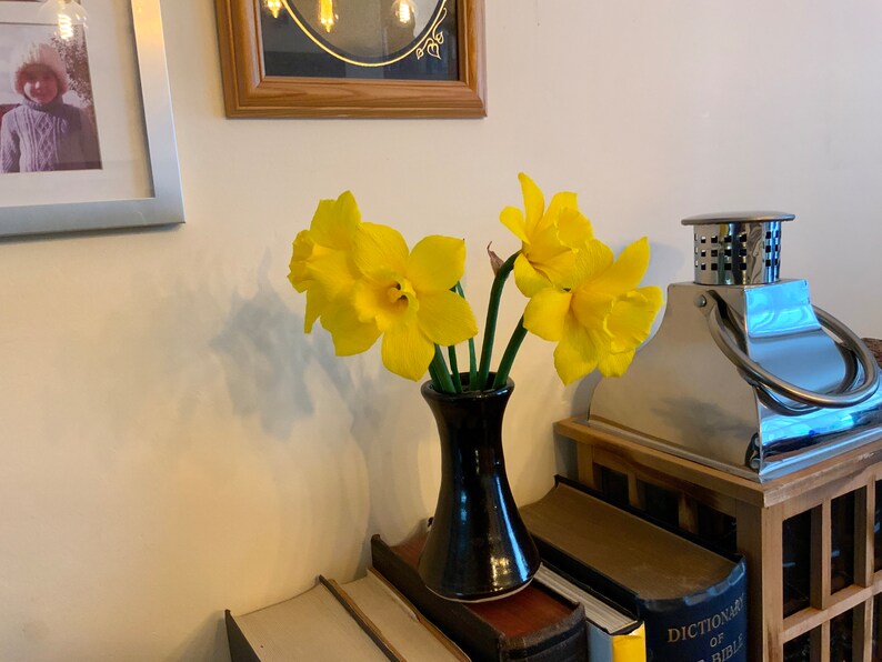 May include: A black ceramic vase holds several bright yellow daffodils. The vase sits atop a stack of books, with a framed photo and a decorative metal lantern in the background. The flowers are in full bloom.