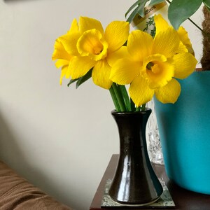 May include: A bouquet of bright yellow daffodils in a dark brown vase. The flowers have textured petals and green stems. The vase sits on a small table next to a turquoise planter with a green plant.