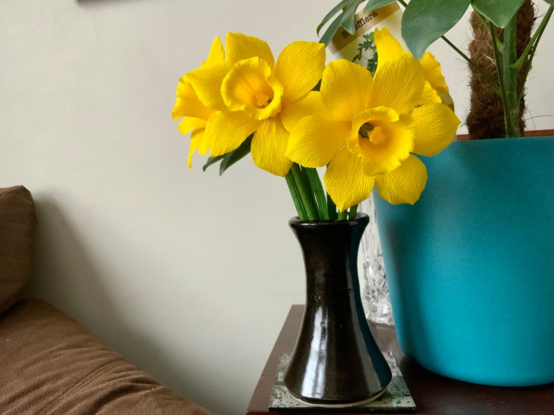 May include: A bouquet of bright yellow daffodils in a dark brown vase. The flowers have textured petals and green stems. The vase sits on a small table next to a turquoise planter with a green plant.
