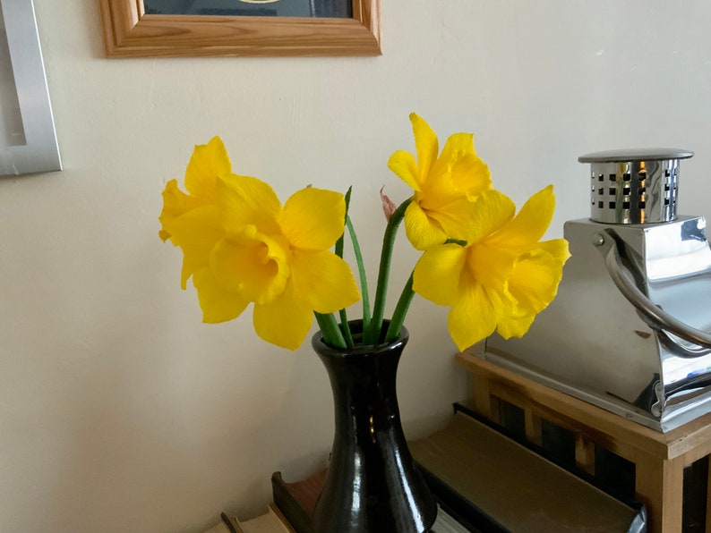 May include: A black vase holds a small bouquet of bright yellow daffodils. The flowers have ruffled petals and green stems. The vase sits on a wooden surface, with a decorative metal lantern in the background.