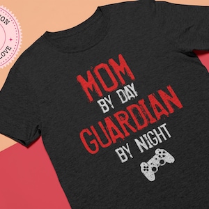 May include: A dark gray t-shirt with red text that reads "Mom by Day Guardian by Night" and a white video game controller graphic.