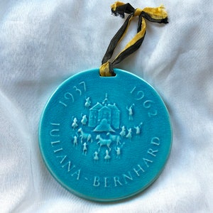 May include: A round, turquoise ceramic ornament with the text "1937" and "1962". The ornament features a raised relief design of a building, people, and animals. It is strung with a black and gold ribbon.