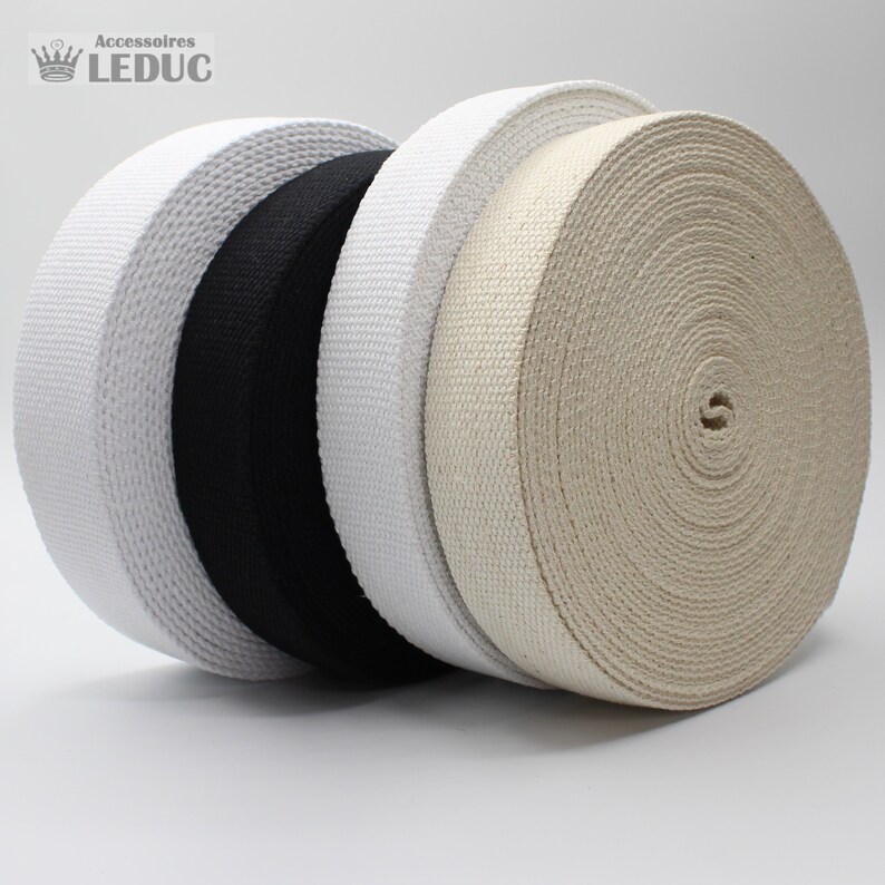 May include: Four rolls of woven webbing in white, black, off-white, and beige. The rolls are tightly wound, showing the texture of the material. The image is well-lit, highlighting the different colors and textures of the webbing.