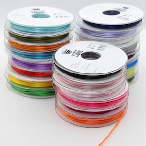 Può includere: Una pila di bobine di nastro di raso colorato. Le bobine sono disposte a forma di arcobaleno, ogni bobina contiene un colore di nastro diverso. Il nastro è largo 3 mm ed è realizzato in 100% poliestere. Le bobine sono etichettate con il nome del marchio "Accessories LEDUC" e il testo "Made in Italy".