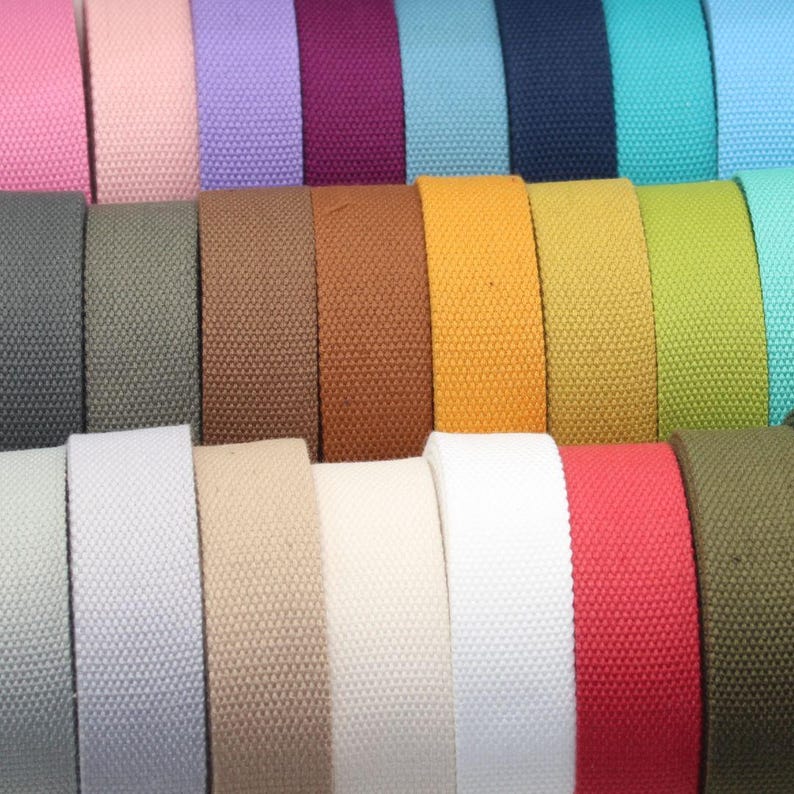 May include: A close-up of a variety of colorful canvas webbing rolls. The colors include pink, lavender, teal, navy, and olive green. Other colors include brown, mustard yellow, white, and red. The webbing is tightly woven.