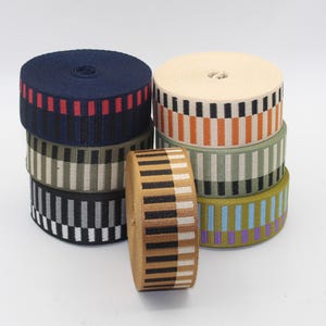 Puede incluir: Una colección de rollos de cinta estampada de colores. Las cintas presentan varios diseños, incluyendo rayas y patrones geométricos en tonos de azul, rojo, negro, blanco, naranja, verde y marrón. Los rollos están apilados y organizados, mostrando la variedad de colores y diseños.