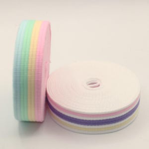 Puede incluir: Dos rollos de cinta de colores pastel. Un rollo está en posición vertical, mostrando rayas de azul claro, amarillo, rosa y verde. El otro rollo está plano, mostrando blanco con rayas rosas, moradas y amarillas. La cinta probablemente se usa para manualidades o costura.