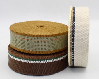 Könnte beinhalten: Drei Rollen gewebtes Gurtband in verschiedenen Farben. Eine Rolle ist beige mit einem grünen und orangefarbenen Streifen. Eine andere ist braun mit einem weißen und grünen Streifen. Die dritte Rolle ist cremefarben mit blauen und braunen Streifen. Das Gurtband ist dicht gewebt.