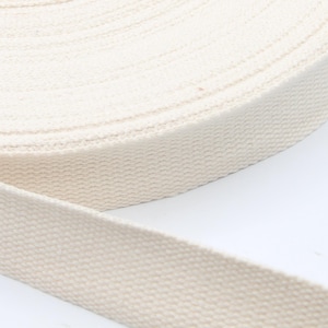 May include: A roll of natural-colored cotton webbing. The webbing is tightly woven and appears to be unwound from a larger roll. The color is a creamy off-white, and the texture is slightly ribbed.