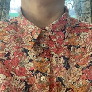 Puede incluir: Camisa abotonada con un vibrante estampado floral en tonos rosa, naranja y crema sobre un fondo azul oscuro. La camisa presenta un cuello cl&aacute;sico y dos botones visibles.