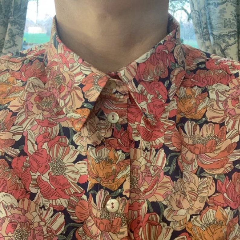 Puede incluir: Camisa abotonada con un vibrante estampado floral en tonos rosa, naranja y crema sobre un fondo azul oscuro. La camisa presenta un cuello cl&aacute;sico y dos botones visibles.