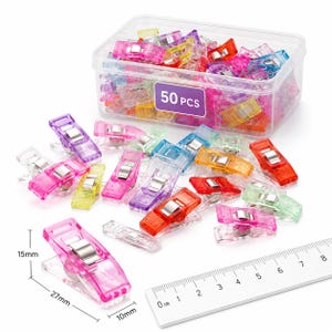 Peut inclure: Un ensemble de 50 pinces à couture en plastique translucide multicolore avec ressorts métalliques, rangées dans une boîte rectangulaire transparente. Une pince rose est présentée près d'une règle, indiquant 27mm de long, 10mm de large et 15mm de haut.