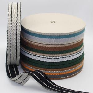 May include: A stack of woven webbing rolls in various colors, including cream, teal, gray, brown, green, and black. One roll is unrolled, displaying a black and white striped pattern. The webbing appears to be made of a durable material.