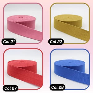 May include: Four spools of colorful webbing ribbon are displayed. The colors include pink (Col 21), gold (Col 22), red (Col 27), and blue (Col 28). Each spool is neatly rolled, with a portion of the ribbon unrolled, showcasing the material's texture.