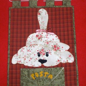 Può includere: Una cassetta della posta in tessuto a quadri rosso e bianco con un'applicazione di cane floreale bianco e una busta verde con la parola "POSTA" ricamata in giallo.