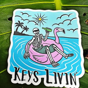 Peut inclure: Un autocollant avec un squelette de dessin animé portant des lunettes de soleil et tenant une bouteille, flottant sur un flotteur de piscine rose en forme de flamant rose dans de l'eau bleue. Le texte "KEYS LIVIN'" est en bas de l'autocollant.