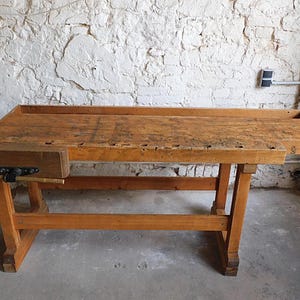 May include: A vintage wooden workbench with a natural finish. The workbench features a large rectangular top with a built-in vise and a lower shelf. The legs and frame are also made of wood. The workbench is approximately 183 cm long.