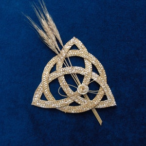 Könnte beinhalten: Ein geflochtener Strohknoten, oder Triquetra, mit einer kleinen weißen Blume in der Mitte, auf blauem Hintergrund.  Weizenhalme sind hinter dem Knoten platziert.