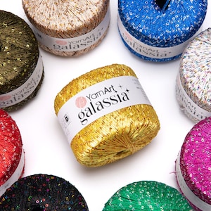 May include: A selection of vibrant yarn skeins in gold, blue, green, and red, with a shimmering, sequined finish. Each skein features a white label with the text "YarnArt + galassia". The yarn is displayed on a white background.
