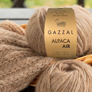 May include: A skein of Gazal Alpaca Air yarn in a light brown color. The yarn is made of 100% baby alpaca wool and is 50 grams per skein. The yarn is shown next to a knitted scarf and a sweater.