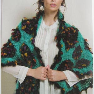 May include: A woman wearing a teal and black crocheted shawl with colorful accents. The shawl has a fringed edge and is draped over a white blouse.