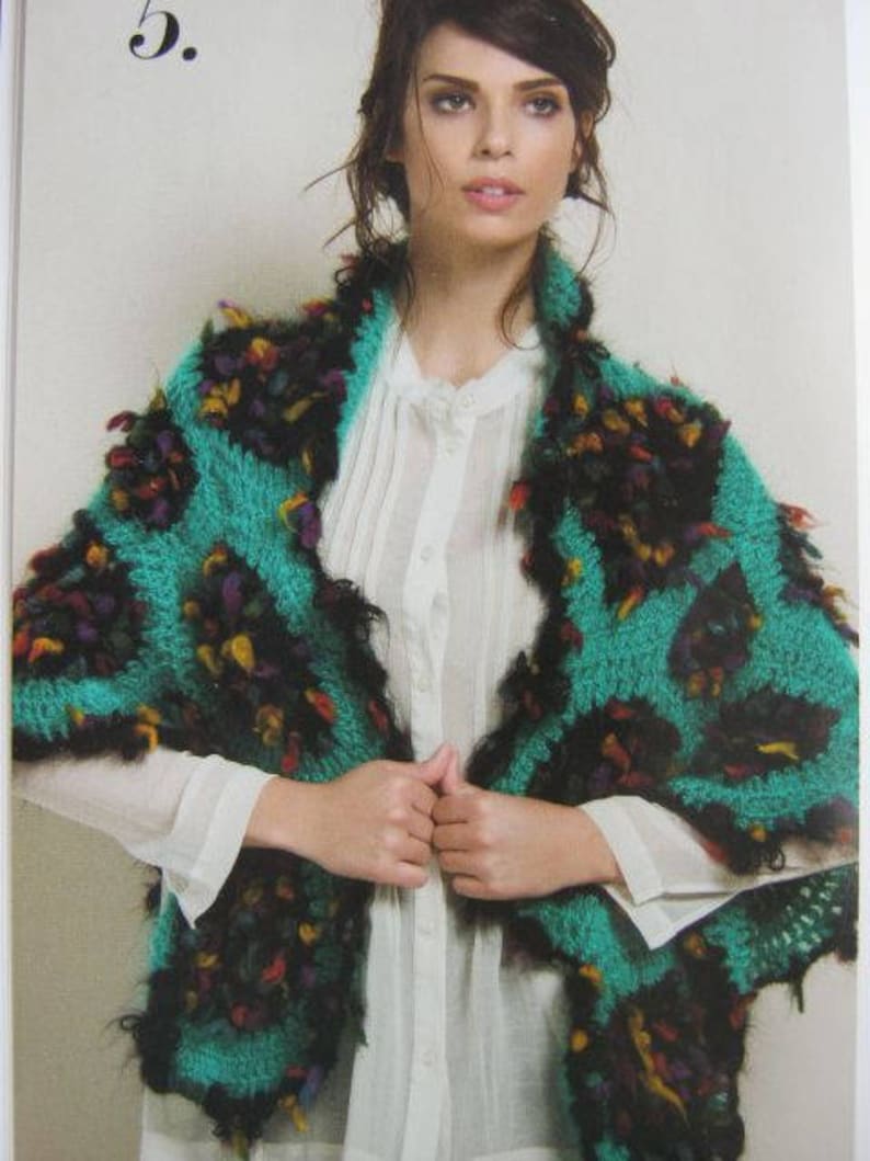May include: A woman wearing a teal and black crocheted shawl with colorful accents. The shawl has a fringed edge and is draped over a white blouse.