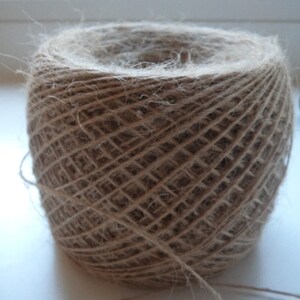 May include: A ball of natural twine, made from jute fibers, is wound tightly in a circular shape. The twine is a light brown color and has a rough texture.