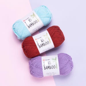 May include: Three skeins of YarnArt Jeans Bamboo yarn in light blue, dark red, and light purple. Each skein features a white paper label with the text YarnArt Jeans Bamboo and a bamboo illustration, noting 50 grams and 150 meters per unit.