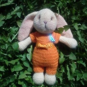 May include: A crocheted bunny toy wearing an orange jumpsuit with a yellow bow and a blue butterfly pin. The bunny is sitting on a bed of green leaves.