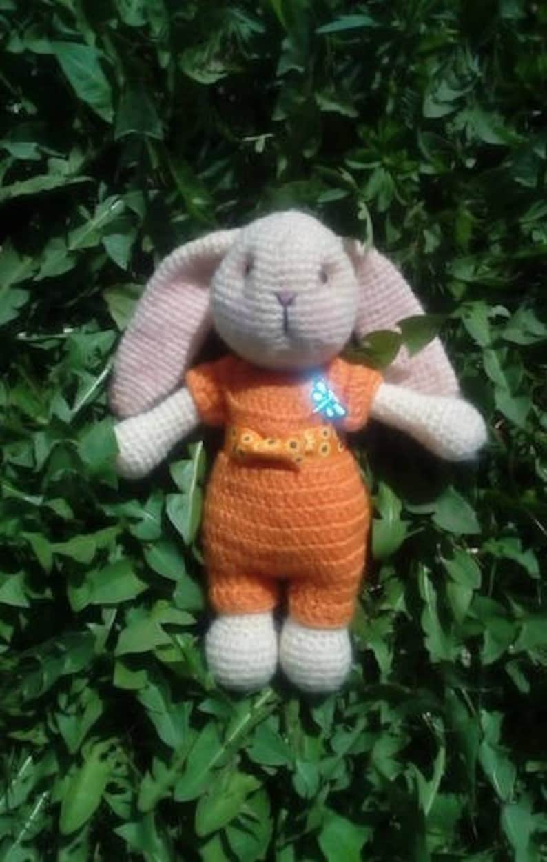 May include: A crocheted bunny toy wearing an orange jumpsuit with a yellow bow and a blue butterfly pin. The bunny is sitting on a bed of green leaves.