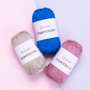 May include: Three skeins of YarnArt macrame cord in blue, beige, and pink. Each cylindrical skein features a white paper label with the brand name YarnArt and the word macrame printed in black text, wrapped around the center of the cord.