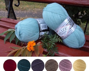 May include: Two balls of light blue yarn on a wooden bench. The yarn is labelled "Alize Superlana" and is a soft, lightweight yarn. The yarn is surrounded by green leaves and a single orange flower.