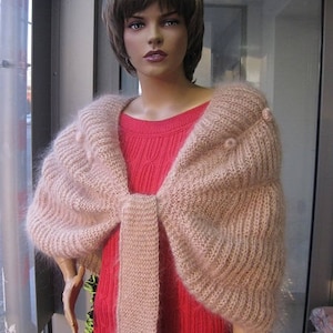 May include: A soft, fuzzy, light pink knitted shrug with a ribbed texture and small pom-pom accents, worn over a bright red sleeveless dress with a vertical cable knit pattern. The shrug features a wide, draped collar and a central tie closure.