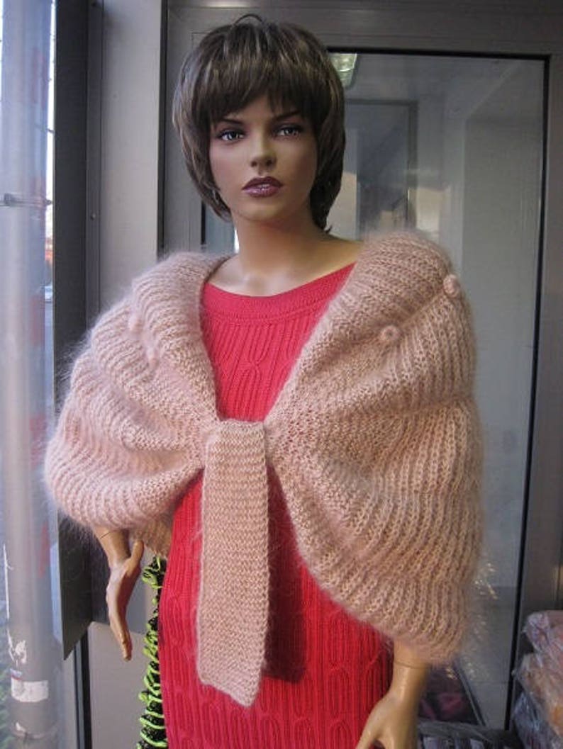 May include: A soft, fuzzy, light pink knitted shrug with a ribbed texture and small pom-pom accents, worn over a bright red sleeveless dress with a vertical cable knit pattern. The shrug features a wide, draped collar and a central tie closure.